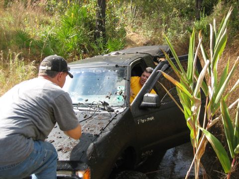 1989 Ford Bronco - A black off-road vehicle is stuck in the mud, with one person in the driver's seat and another person outside the hood.