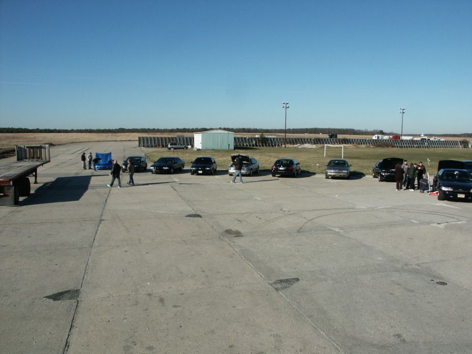 Several cars are parked on a concrete lot with a few people standing around. A trailer is on the left