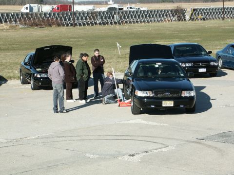 2001 Mercury Grand Marquis - A group of people are gathered around a black sedan that is being jacked up, with another similar car parked nearby.