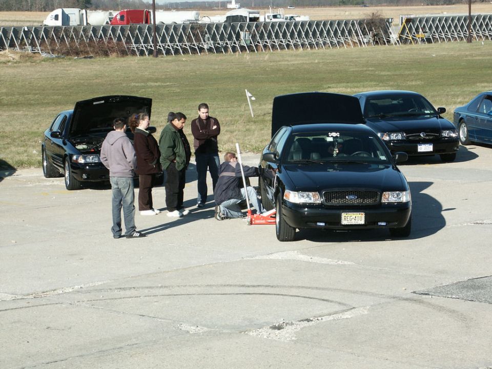 people gathered around a black sedan, one person is using a jack on the front wheel. Another black sedan is parked behind it