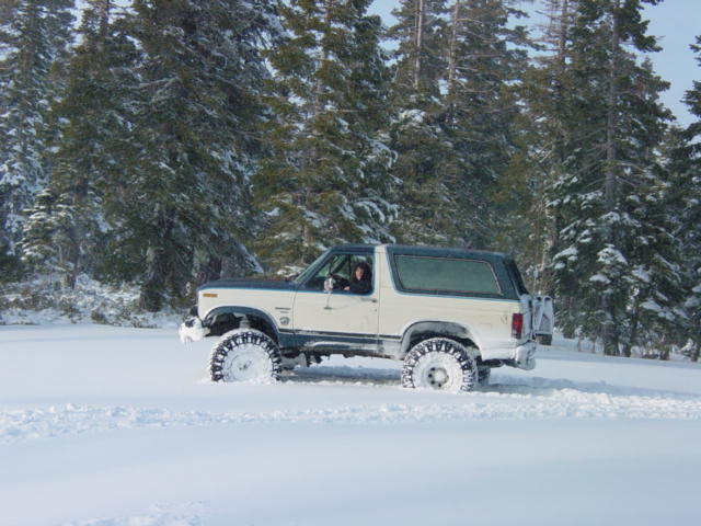 White Ford Bronco with dark top driving in deep snow, trees in background