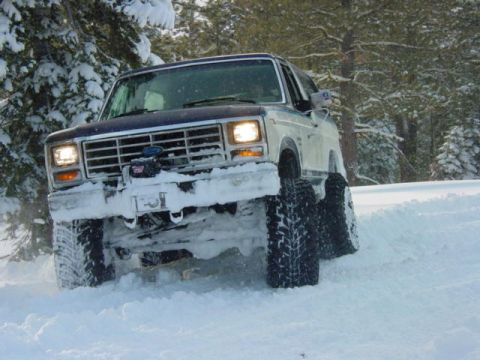1986 Ford Bronco - This lifted 1986 Ford Bronco navigates a snowy forest road, its large tires kicking up snow.