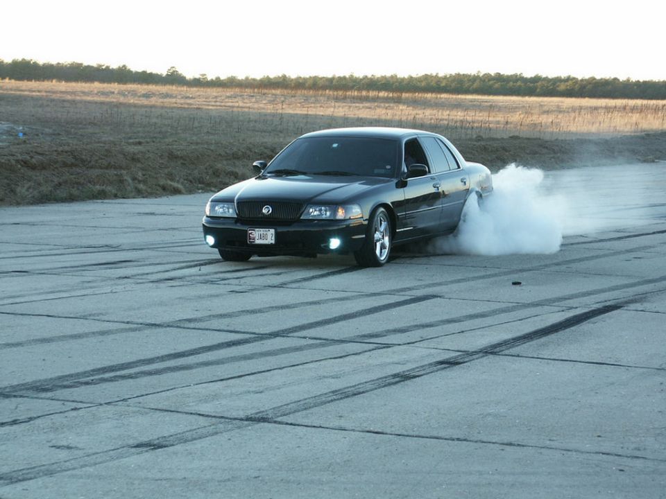 Black sedan with aftermarket grille and wheels doing a burnout, emitting smoke from rear tires