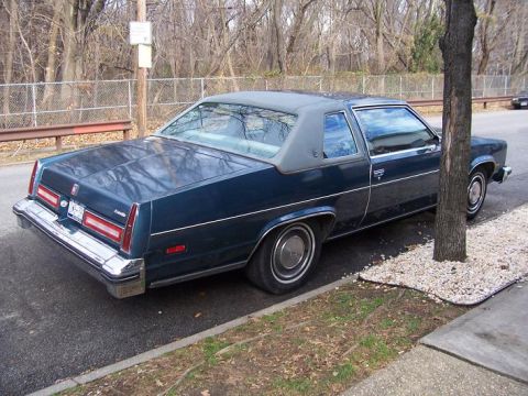 1999 Ford Crown Victoria - A dark blue 1977 Oldsmobile Delta 88 Royale coupe is parked curbside, showing its rear 3/4 profile.