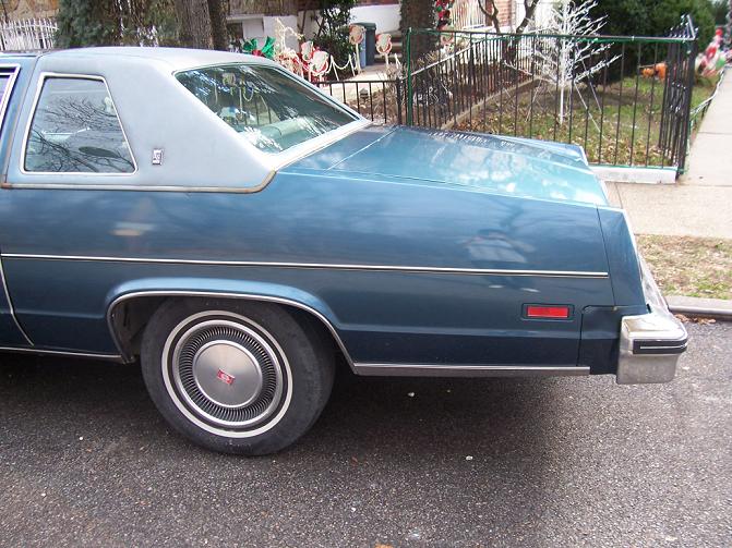 Rear quarter view of a blue sedan with a light-colored vinyl roof and chrome trim. It has a red reflector on the rear fender