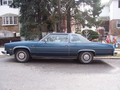 1999 Ford Crown Victoria - This blue coupe, likely a 1977 model, is parked curbside in front of residential homes.