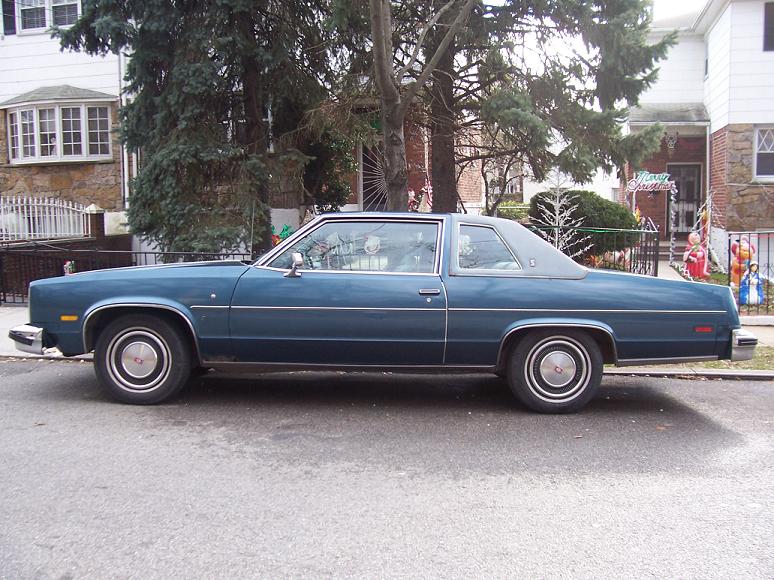 Blue coupe car parked on street, side view. Appears to be from the late 1970s