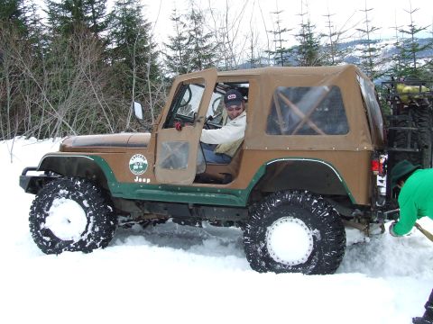 1976 Ford F-150 - A brown Jeep Wrangler with a tan soft top is stuck in the snow, with a person in the driver's seat and another attempting to free it.