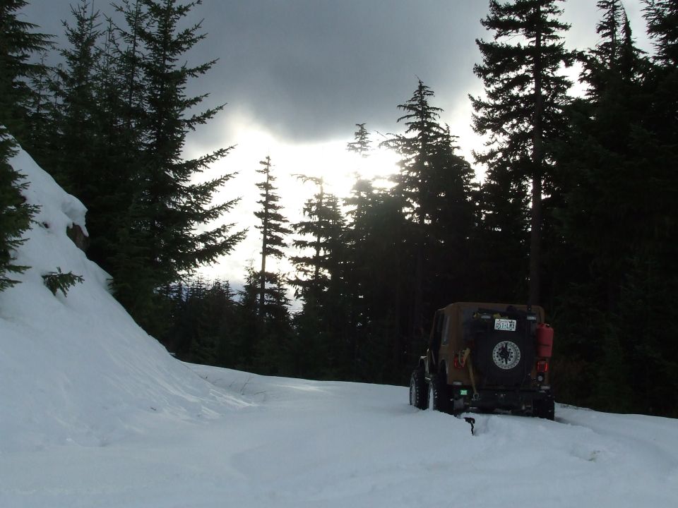 Jeep Wrangler on snowy road in forest, sun breaking through clouds