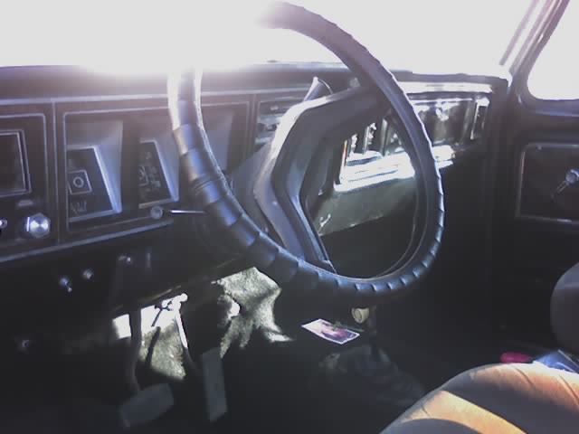 Sunlit interior of a Ford Bronco showing black steering wheel, dash, floor shifter, and striped seat.