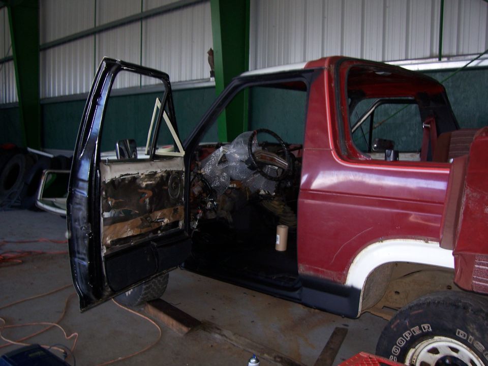 Red Ford Bronco in a shop with driver's door open, stripped interior and door panel during restoration