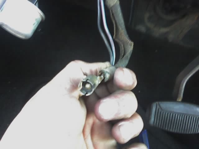 Hand holding a wiring harness connector with wires. Vehicle interior with pedals visible