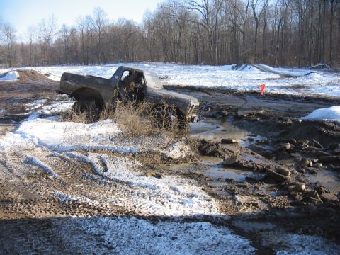 1979 Ford Bronco - A pickup truck is stuck in a muddy ditch with snow and trees in the background.
