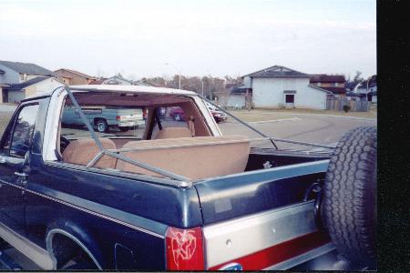 1990 Ford Bronco - Rear view of a 1990 Ford Bronco showing the interior and a spare tire. Another vehicle is parked behind it.