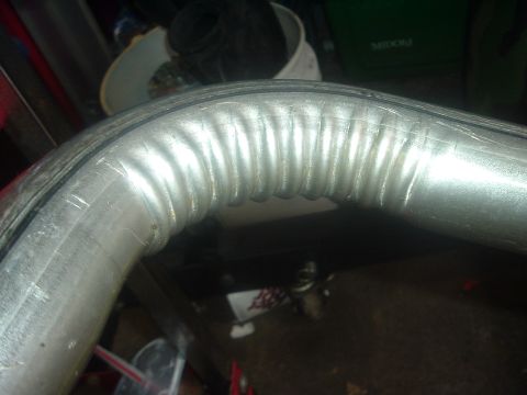 1989 Ford Escort - A close-up view of a bent metal exhaust pipe, showing its ribbed texture and a black seal.