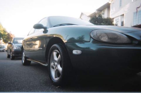 1999 Mercury Sable - A dark green sedan is parked on a street, with other vehicles visible in the background. The image includes a watermark for SUPERMOTORS.