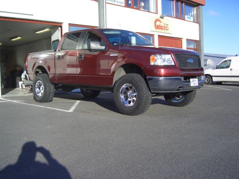1996 Ford Bronco - This red Ford F-150 pickup truck features a lifted suspension and chrome wheels, parked in front of a commercial building.