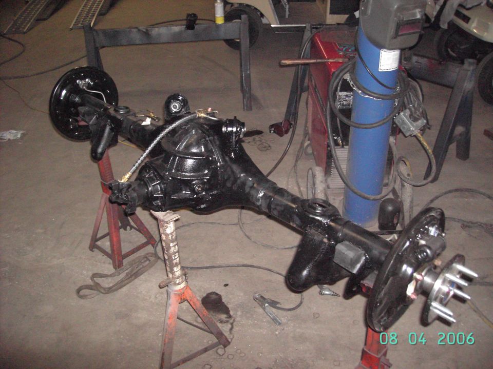 Black-painted rear axle assembly on red jack stands in a garage, welder in background, dated 08 04 2006