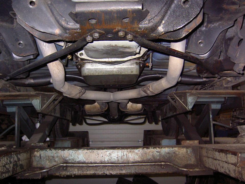 Underbody view of a car on a lift showing oil pan, crossmember, and dual exhaust pipes merging into a Y-pipe.