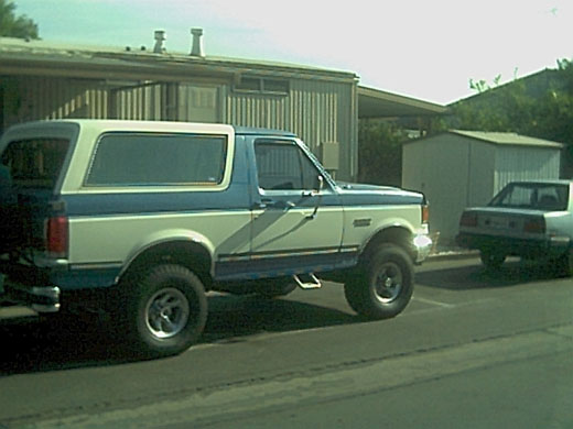 Blue and white Ford Bronco with lift and chrome wheels, side view, parked on a street