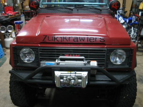 1987 Suzuki Samurai - Front view of a red off-road vehicle with custom text on the hood and a winch mounted on the bumper.