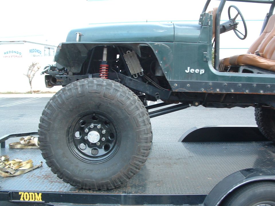 Green Jeep with large tires on a trailer, front fender removed, showing suspension