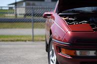 1991 Ford Probe - Close, low front-corner view of a dark red sports coupe with its hood raised, showing the pop-up headlamp housing, amber corner marker lens…