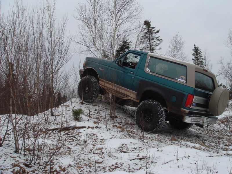 Lifted teal and tan Ford Bronco on large off-road tires climbing a snowy mound in winter
