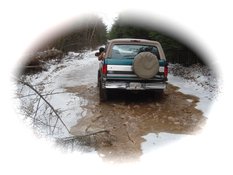 Rear view of teal and tan Ford Bronco with spare tire cover on a muddy snowy trail