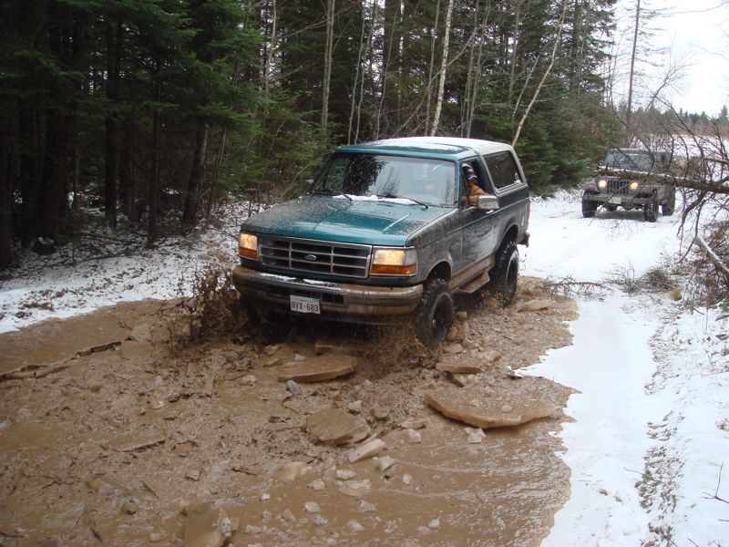 Teal Ford Bronco with white hardtop splashing through muddy puddle on a snowy trail, Jeep behind