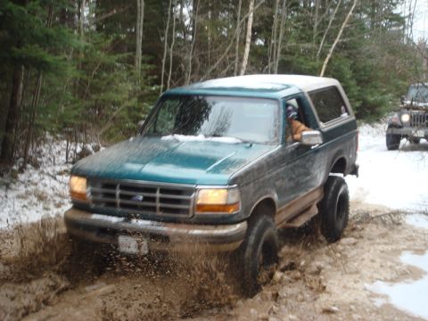 1996 Ford Bronco - A 1996 Ford Bronco navigates a muddy trail in a snowy forest, with another vehicle visible in the background.