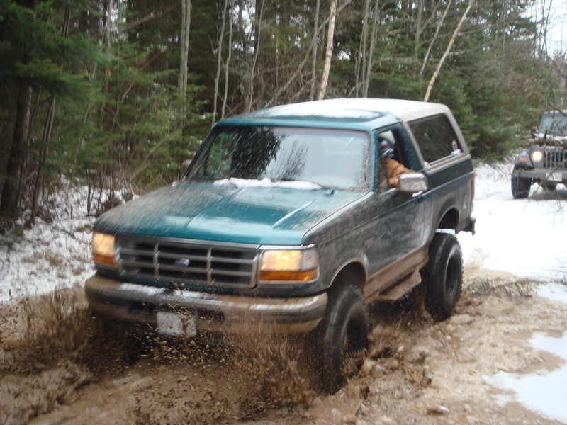 Teal Ford Bronco with white hardtop driving through mud on a snowy woods trail, driver at window