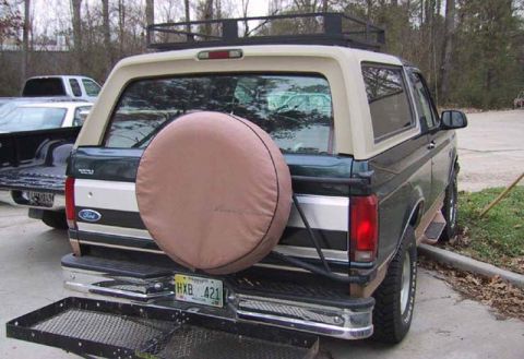 1995 Ford Bronco - The rear of a 1995 Ford Bronco is shown, a spare tire cover and a roof rack, with another vehicle beside it.
