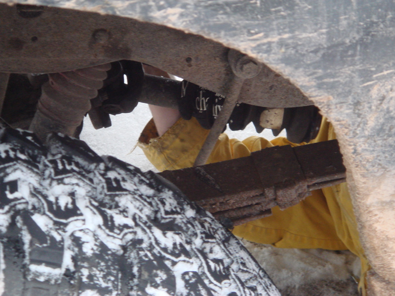 Inside a wheel well showing rusty leaf spring pack and coil spring behind a snowy mud tire