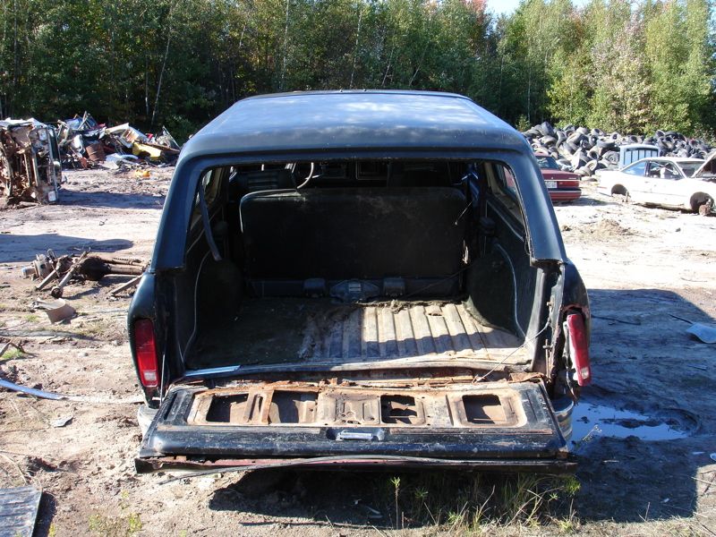 Back of an old SUV, tailgate down, showing empty cargo area. Junkyard in background with other vehicles and scrap