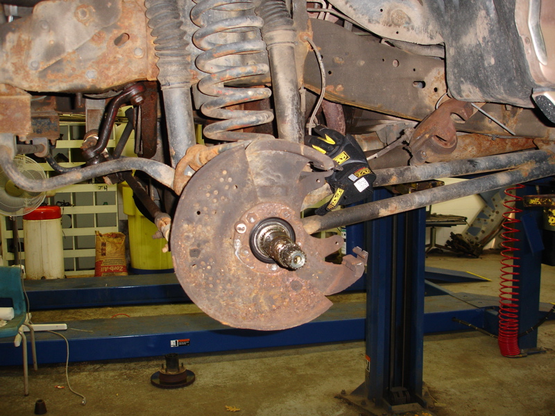 Rusty brake rotor and suspension components on a vehicle lift. A gloved hand is visible