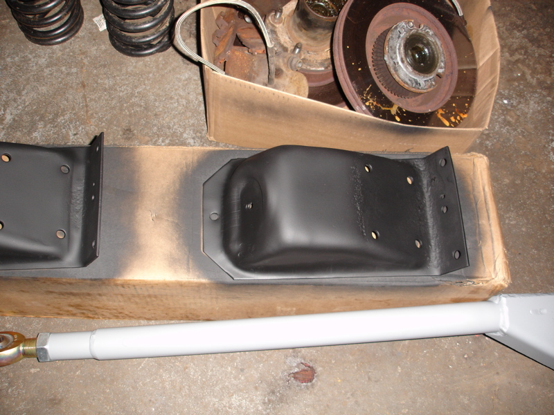 Black painted suspension brackets and a white adjustable control arm sit on a cardboard box. Springs and a brake rotor are in the background