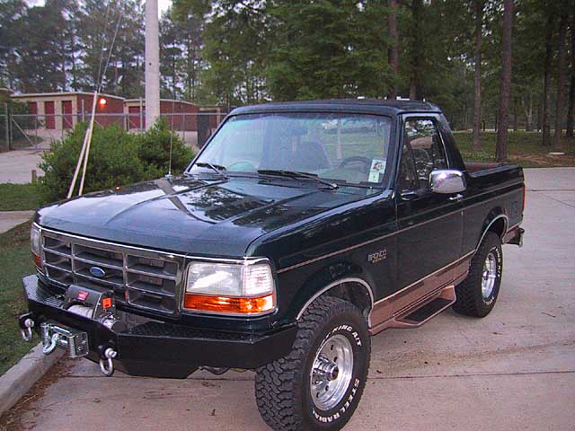 Dark green Ford Bronco with topless rear, winch bumper and all-terrain tires on a driveway