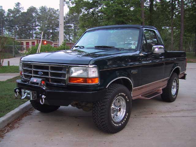 Dark green Ford Bronco with winch bumper and alloys, front 3/4 view on driveway
