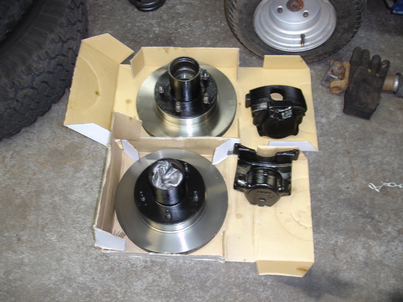 New brake rotors and calipers laid out in cardboard boxes on a concrete floor. A tire and a glove are visible in the background