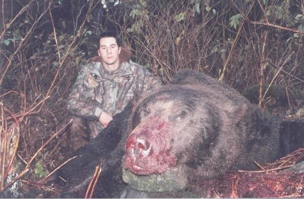 Hunter in camouflage poses next to a large dead bear in a wooded area