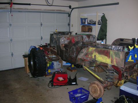 1986 Ford Bronco - A vehicle chassis is undergoing restoration in a garage, with welding equipment nearby and a large tire leaning against a workbench.