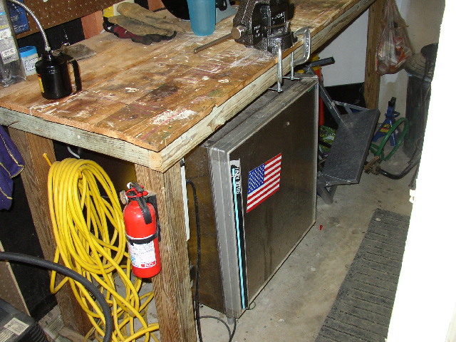 Workbench with a vise, oil can, yellow extension cord, and red fire extinguisher; a mini-fridge with an American flag sticker is below the workbench