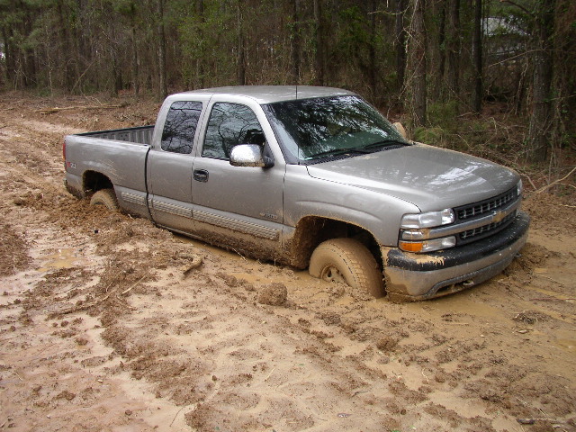 Silver pickup truck stuck in mud, front 3/4 view. Mud covers the tires and lower body