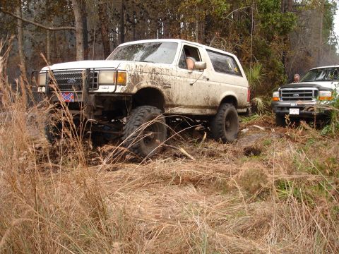 1989 Ford Bronco - A lifted white 1987 Ford Bronco appears stuck in the mud while another truck waits behind it in a wooded area.