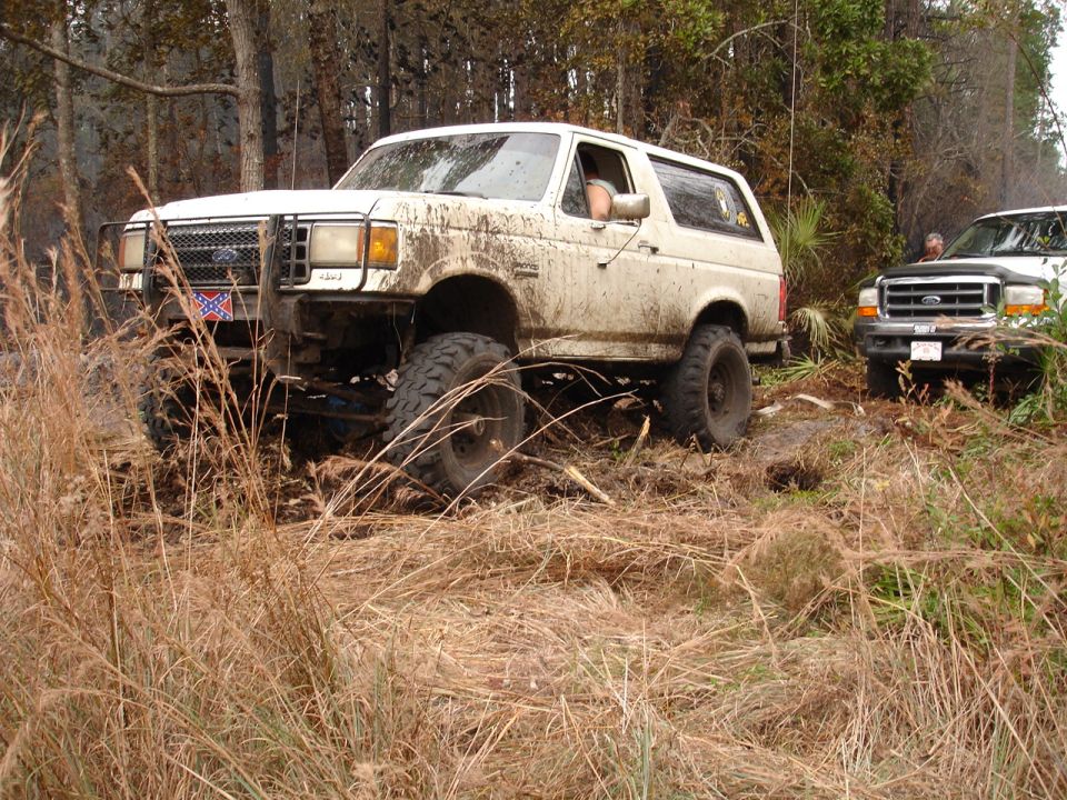 White 1987 Ford Bronco with lifted suspension stuck in muddy terrain, another truck visible behind