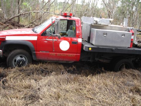 1989 Ford Bronco - A red fire department truck is stuck in a muddy, grassy area with trees in the background.