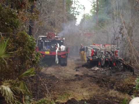 1989 Ford Bronco - Firefighters are working in a wooded area with two fire trucks and smoke visible.