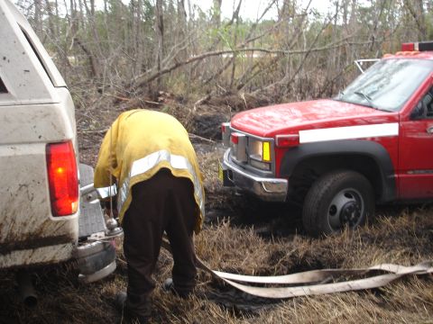 1989 Ford Bronco - A firefighter connects a hose between a white SUV and a red truck in a muddy, wooded area.