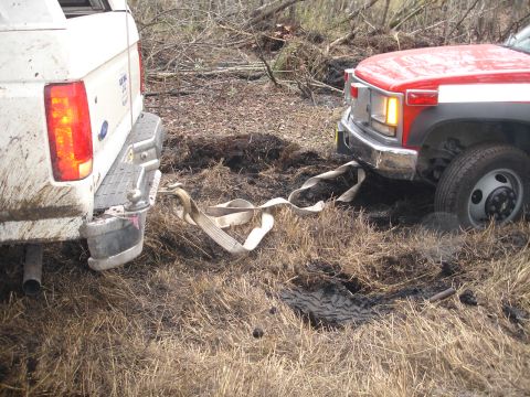 1989 Ford Bronco - A white 1989 Ford Bronco and a red truck are stuck in muddy terrain, with a tow strap connecting them.